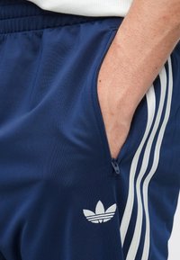 Calças de jogging atléticas sintéticas azul-marinho com três riscas brancas na lateral e um bolso com fecho. Apresentam um pequeno emblema com logótipo.