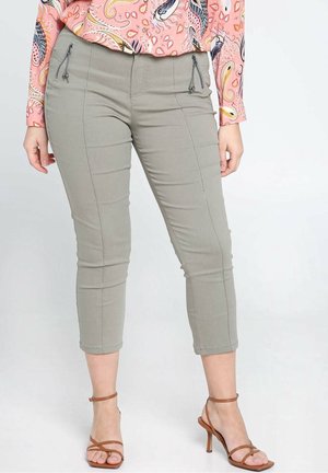 Frau trägt hellgraue kurze Hosen mit Reißverschlusstaschen, eine pinke Paisley-Bluse und braune Sandaletten mit Absatz, steht vor einem weißen Hintergrund.