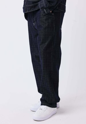 Personne portant un pantalon foncé à motifs et des baskets blanches, debout avec une main dans la poche sur un fond clair uni.