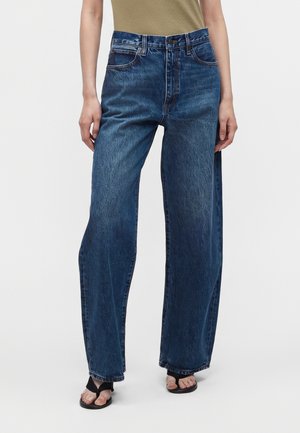 Femme portant un jean denim bleu à jambes larges avec poches avant, associé à des sandales noires ouvertes à l’avant et un haut beige.