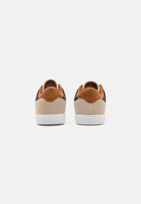 Pair of low-top sneakers viewed from the back, featuring beige suede, brown leather heel tabs, black side panels, and white rubber soles.