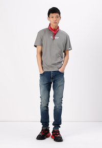 Camiseta de algodón gris con estampado de logo, jeans de mezclilla azul y zapatillas negras con acentos rojos. Se lleva una bandana roja anudada alrededor del cuello.