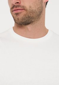 Κοντινό πλάνο του κάτω μέρους του προσώπου ενός άνδρα με ελαφρύ μούσι και μουστάκι, που φοράει ένα απλό λευκό μπλουζάκι με στρογγυλή λαιμόκοψη.