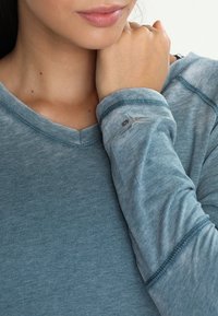 Bluzka z długim rękawem i dekoltem w kształcie litery V wykonana z miękkiej, melanżowej niebieskiej tkaniny o subtelnej fakturze i wzmocnionych szwach; posiada logo na rękawie.