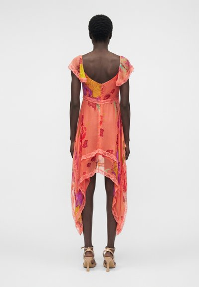 Γυναίκα φοράει αμάνικο, ασύμμετρο κοραλλί φόρεμα με λουλουδένια σχέδια, δαντέλα και πέδιλα με ψηλό τακούνι και λουράκι στον αστράγαλο, με την πλάτη γυρισμένη.