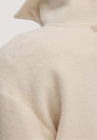 Abrigo beige de mezcla de lana con un acabado suave y texturizado, con cuello de solapa y una silueta relajada. Vista en primer plano de la tela.