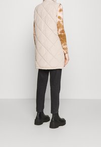 Gilet smanicato beige trapuntato con motivo a diamante, abbinato a una maglietta a maniche lunghe marrone tie-dye, jeans neri e stivali.