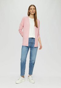 Lichtroze cardigan met open voorkant, lange mouwen en zachte textuur, gecombineerd met een wit T-shirt en relaxed-fit blauwe spijkerbroeken, en witte sneakers.