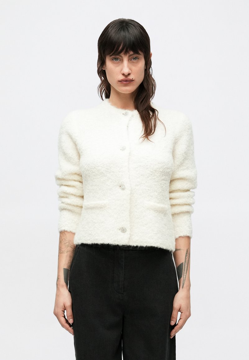 Bela, puhasta jopica z okrolim izrezom, šestimi dekorativnimi gumbi in dvema sprednjima žepoma. Ohlapen kroj z manšetnimi rokavi.