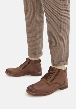 Brūnas ādas potīšu zābaki ar apaļu purngalu, priekšējo šņorēšanas daļu un sānu rāvējslēdzēju. Teksturēta virsma un kontrastējoša sarkanā apdare uz zolēm.