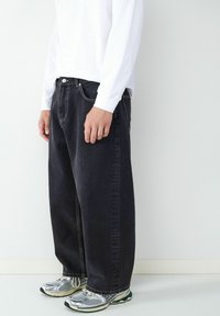 Czarne dżinsy o luźnym kroju, widoczne detale ściegów, noszone z białą koszulą z długim rękawem i srebrnymi butami do biegania.