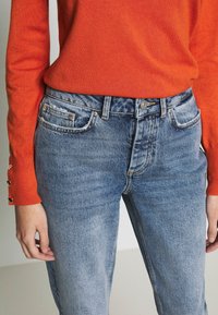Orange knitted sweater with a round neckline and buttoned cuffs, paired with mid-blue faded jeans featuring a classic five-pocket design.