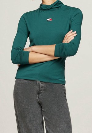 Person iført en teal langærmet top med en lille Tommy Jeans-logo, armene krydset, kombineret med højtaljede grå denimjeans.