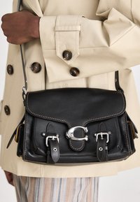 Crna kožna crossbody torba s crnim metalnim detaljima, dva kopča i kontrastnim bež šavovima. Nosena uz svjetlosivu bež kaput.