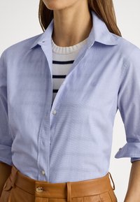 Lys blå skjorte med knappelukking og oppkvelte ermer, myk tekstur og diskret mønster. Brukt over en hvit og marineblå stripete genser.