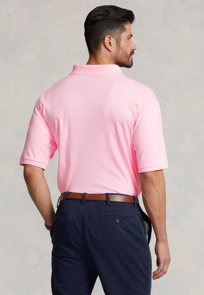 Rožinė polo marškinėliai su standartiniu apykakle ir trumpomis rankovėmis, derinami su tamsiai mėlynomis kelnėmis. Marškinėliai turi lygią tekstūrą ir laisvą siluetą.