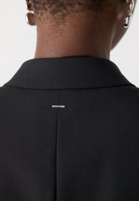 Čierny blazer s golierom, ktorý má viditeľný šev na zadnej strane a akcent striebornou bezpečnostnou ihlou. Hladká textúra, štruktúrovaný dizajn, minimalistické detaily.