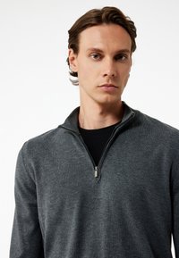 Harmaa vetoketjullinen villapaita, jossa on korkea kaulus ja sileä pinta. Siinä on metallivetoketju ja musta vuori.