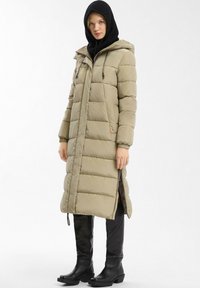 Lange, gevoerde beige jas met een capuchon, voorzien van een rits en trekkoordaccenten. Draag het over een zwarte trui, gecombineerd met kniehoge zwarte laarzen.