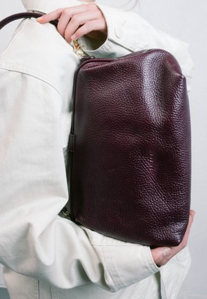 Persona con chaqueta clara sostiene un bolso de hombro de cuero granate oscuro con textura, con cierre dorado junto al hombro y la parte inferior.