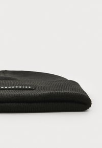 Black knitted beanie made from ribbed fabric, featuring a flat folded design and a rectangular brand label stitched on the front.