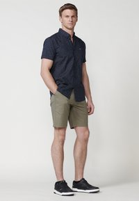 Majica kratkih rukava u tamnoplavoj boji s točkastim uzorkom, uparena s maslinasto zelenim šortsevima. Model nosi crne tene. Jednostavan, ležeran dizajn.