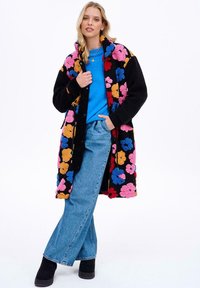 Farebný kabát s kvetinovým vzorom a čiernym materiálom, s fleecovým povrchom, prednými vreckami, nosený cez modré tričko a spárovaný so svetlomodrými džínsy a čiernymi čižmami.