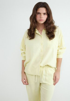 Mladá žena s dlhými hnedými vlasmi, ktorá má na sebe voľnú bledožltú košeľu na gombíky a zodpovedajúce nohavice so sťahovacou šnúrkou, stojaca pred svetlosivým pozadím.