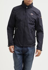 Mörkblå vattentät jacka med hög krage, dragkedja framtill och två sidofickor. Har en logotyp på övre vänster bröst.