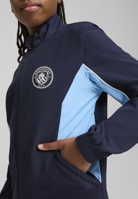 Veste zippée bleu marine et bleu clair avec logo de Manchester City, poignets élastiques et poches latérales au design de panneaux contrastés.