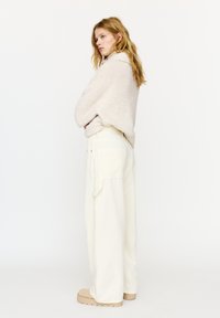 Młoda kobieta stojąca bokiem, ubrana w kremowy, puchaty sweter, szerokie białe spodnie z dużymi kieszeniami i beżowe buty na platformie.