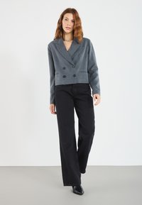 Grå, kort dobbelspent blazer med skjortekrage og to svarte knapper, kombinert med svarte vide jeans og spisse svarte sko.