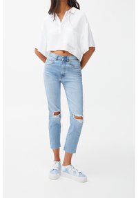 Femme portant un haut court blanc, un jean déchiré taille haute bleu clair et des baskets tie-dye bleues, posant devant un fond blanc uni.