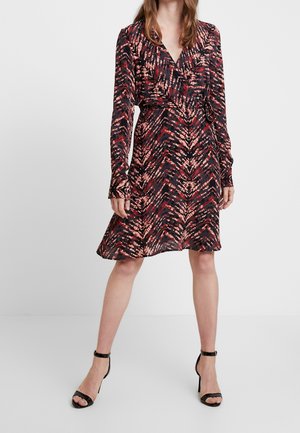 Mujer que lleva un vestido cruzado de manga larga hasta la rodilla con patrones abstractos en rojo, negro y beige, y tacones negros con tiras en el tobillo.