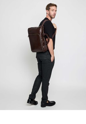 Man wearing black shirt and dark pants carries large brown leather backpack over one shoulder, standing against plain background.