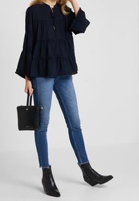 Blouse tiered bleu marine avec des manches évasées, associée à un jean skinny bleu et des bottines noires à la cheville. Sac à main noir porté dans la main gauche.