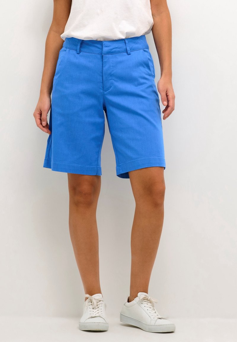 Personne portant un short bleu aux genoux, un t-shirt blanc et des baskets blanches, debout devant un fond blanc uni.