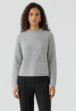 Femme aux cheveux bouclés portant un pull côtelé gris clair avec une petite poche avant et un pantalon noir sur un fond clair uni.