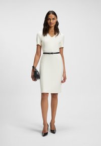 White fitted dress with short sleeves, V-neck, and a narrow black belt. Worn with black pointed-toe heels and a small black handbag.