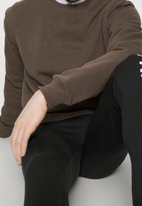 Hnědá mikina se texturovaným logem na hrudi, žebrovanými manžetami a kulatým výstřihem, spárovaná s černými fitted kalhotami.