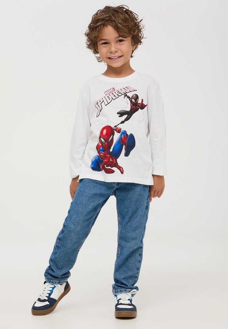 Camicia bianca a maniche lunghe con grafica di Spider-Man, jeans blu e sneakers con accenti bianchi, blu navy e marroni. Il bambino mostra un'espressione allegra.
