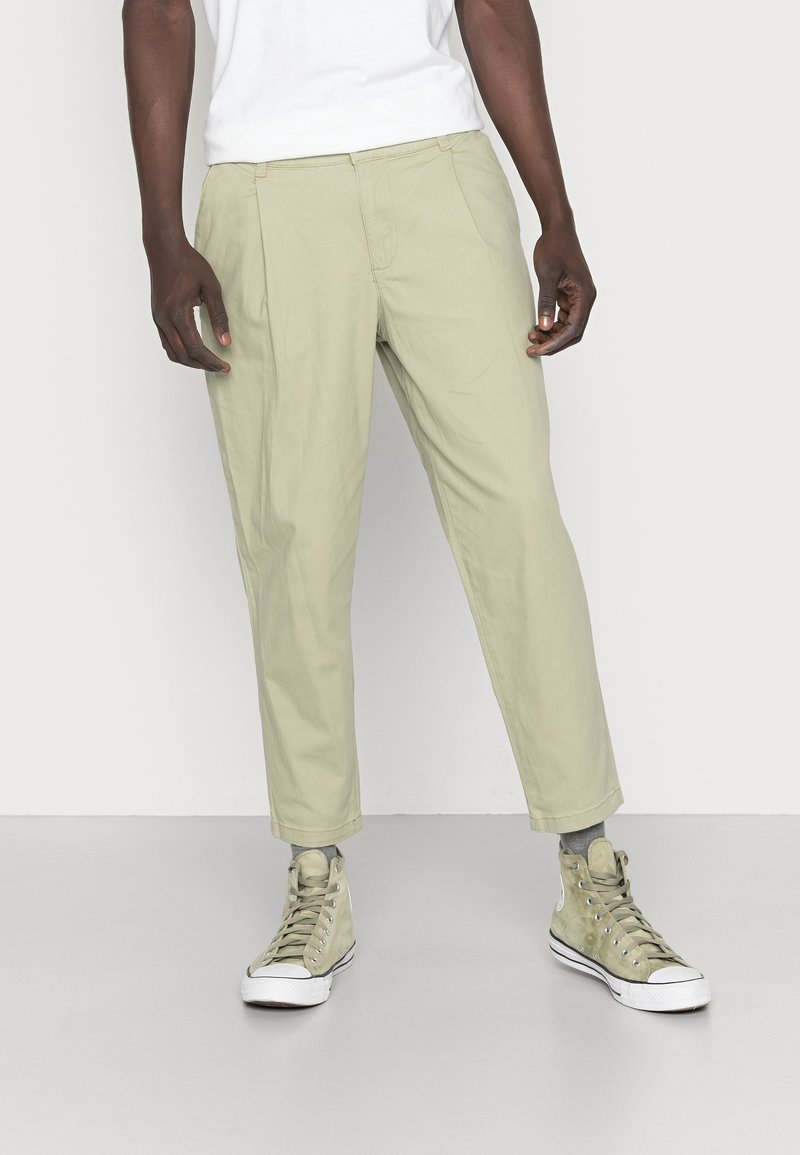 Man med beige kortbyxor, grå strumpor och ljusgröna högsko-sneakers med snören står på grått golv.