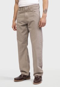 Pantalones de pierna ancha beige en suave sarga de algodón con trabillas, bolsillos frontales y un diseño clásico de cinco bolsillos, combinados con zapatos marrones oscuros.