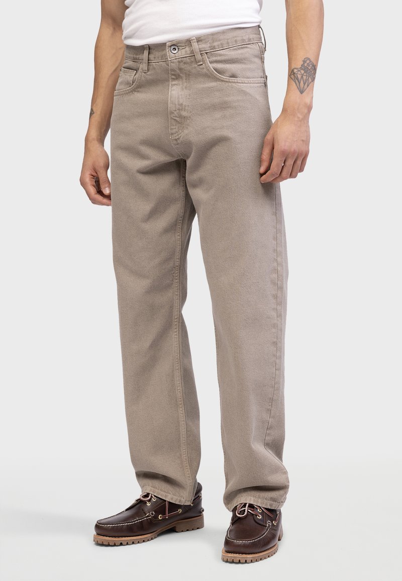 Pantalones de pierna ancha beige en suave sarga de algodón con trabillas, bolsillos frontales y un diseño clásico de cinco bolsillos, combinados con zapatos marrones oscuros.