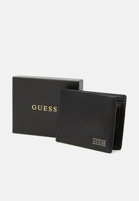 Portefeuille en cuir noir avec plusieurs emplacements pour cartes et une poche zippée, arborant un logo "GUESS" en doré. Accompagné d'une boîte noire.