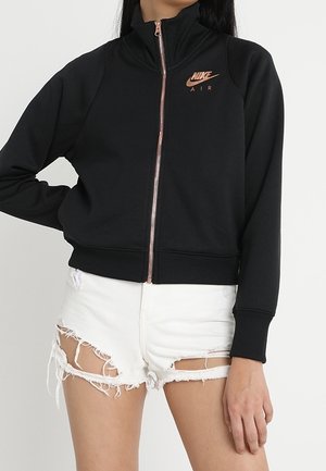 Osoba ubrana w czarną bluza Nike Air na zamek błyskawiczny z różowo-złotym suwakiem i logo, zestawiona z białymi, postrzępionymi szortami jeansowymi.