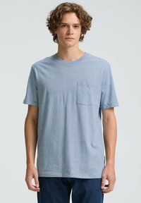 Tricou scurt, cu mânecă scurtă, de culoare albastru deschis, fabricat dintr-un material moale, cu un decolteu rotund și un buzunar pe pieptul stâng. Croi lejer, cu mânecile răsucite.