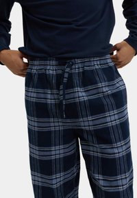Granatowe flanelowe spodnie piżamowe w kratę z regulowanym sznurkiem w talii. Charakteryzują się wzorem w kratkę w odcieniach ciemnego i jasnego niebieskiego. Miękka tekstura tkaniny.