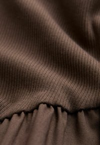 Bruine, ribbelige gebreide stof met variatie in textuur; heeft een geaccentueerd gedeelte aan de onderkant, wat zorgt voor een zachte, vallende uitstraling.