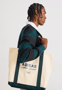 Krēmkrāsas auduma soma ar tumši zaļām siksnām, uz kuras ir melnā krāsā uzraksti "ADIDAS" un "Sporta Apģērbs". Valkāta uz pleca.